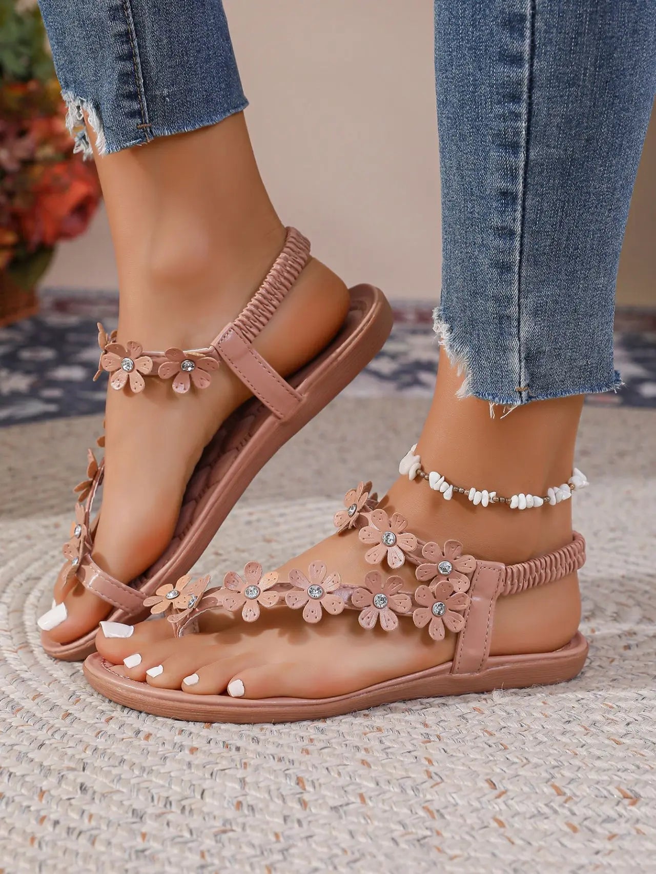 Close-up of pink bohemian flat thong sandals with delicate floral appliques and clear rhinestone embellishments, worn on feet with blue denim jeans and a white beaded anklet.