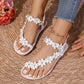 White bohemian flat thong sandals embellished with delicate fabric flowers and sparkling rhinestones, shown on a person's feet paired with blue jeans and a gold anklet.