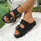 Black denim-inspired slide sandals with frayed edges and gold buckles, styled on feet against a white pebble and green plant background.