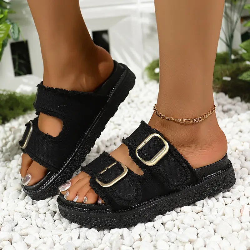 Black Kovan denim-inspired slide sandals with gold buckles and a chunky platform sole, worn on feet with a gold anklet against a white pebble and green foliage background. The distressed denim fabric has frayed edges.