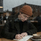 Kovari urban docker beanie in rust orange knit worn by a young man writing in a cafe behind a rainy window.