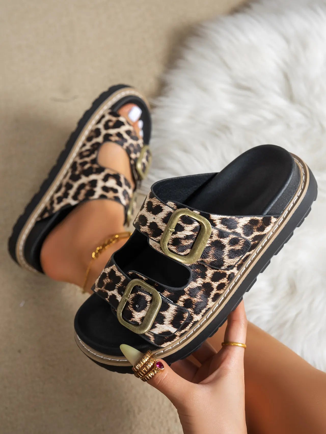 Women's Kovari leopard print platform slide sandals with antique gold double buckle detail, styled on a foot and held by hand against a textured neutral background.