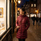 Woman wearing a Kovera burgundy long puffer coat with a slim fit and hood, standing on a cobblestone street at night.