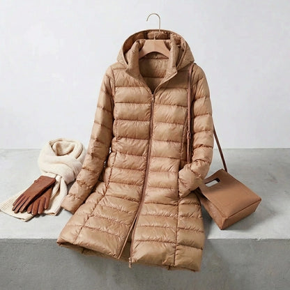 Kovera long puffer coat in khaki with removable hood, styled with a scarf, leather gloves, and a handbag on a studio background.
