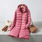 Kovera long puffer coat in soft rose pink with removable hood, styled with a scarf, gloves, and handbag on a concrete surface.