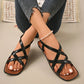 Koyami women's black flat sandals featuring a square toe and criss-cross straps, shown on a model's feet with distressed jeans and a pearl anklet. These strappy summer sandals offer bohemian beach style with comfortable walking design.