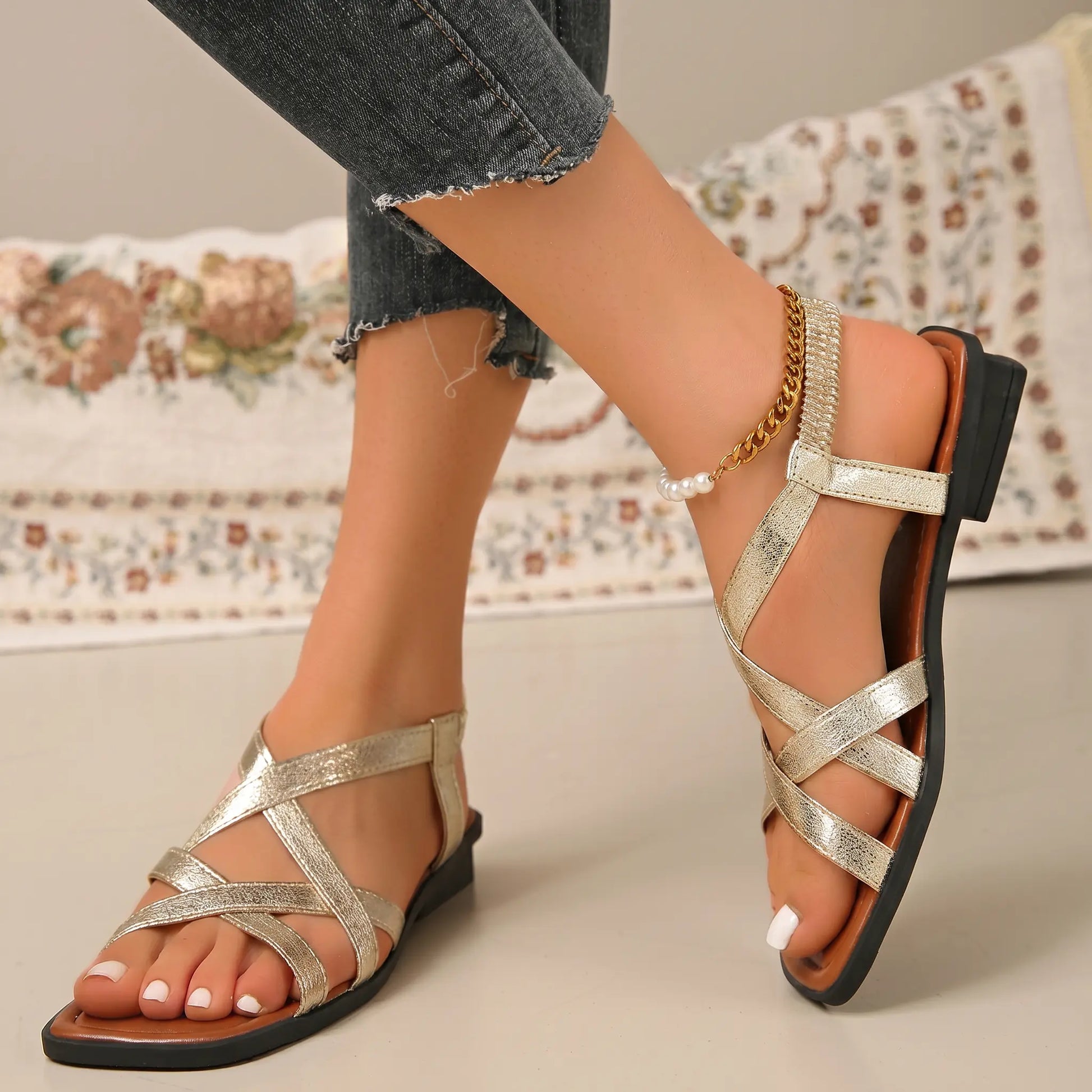 A woman's foot wearing gold strappy flat sandals with a square toe and an elasticated heel strap adorned with a gold chain and pearl accent.