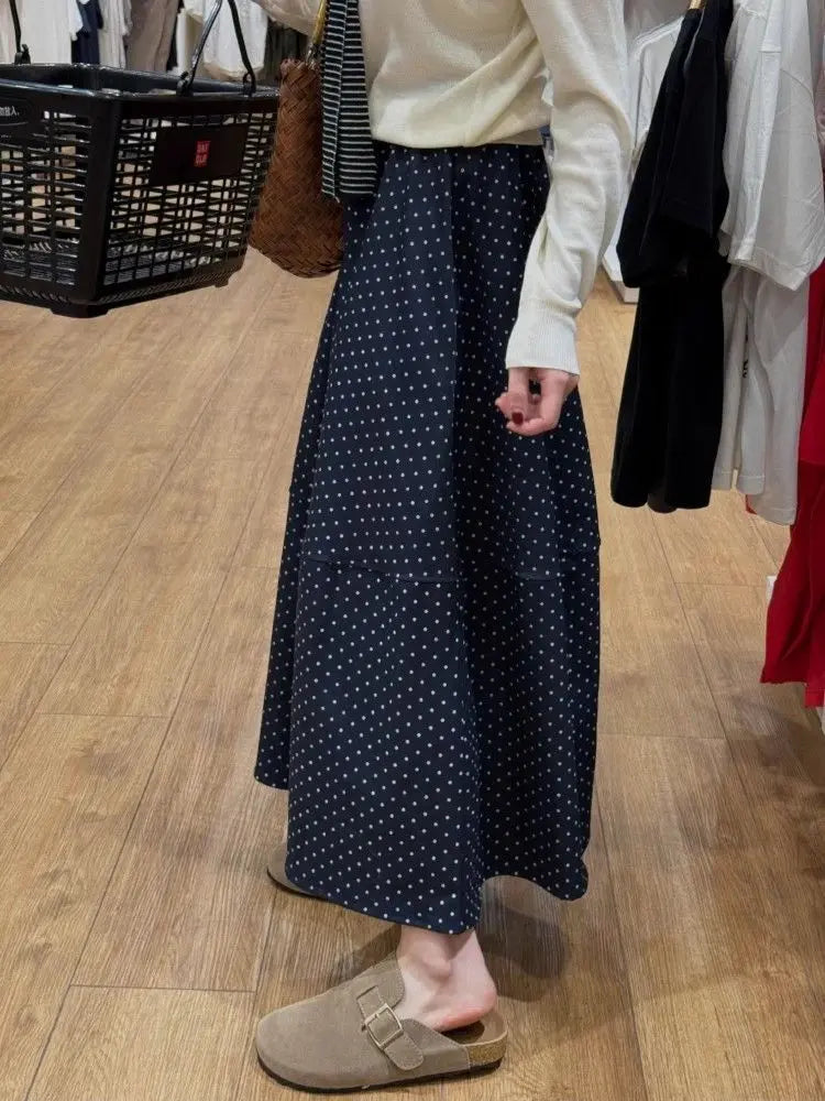 Kozira midnight A-line midi skirt with white polka dot pattern and distinct horizontal panel seam, worn by a model with a white sweater and tan clogs in an elegant indoor setting.