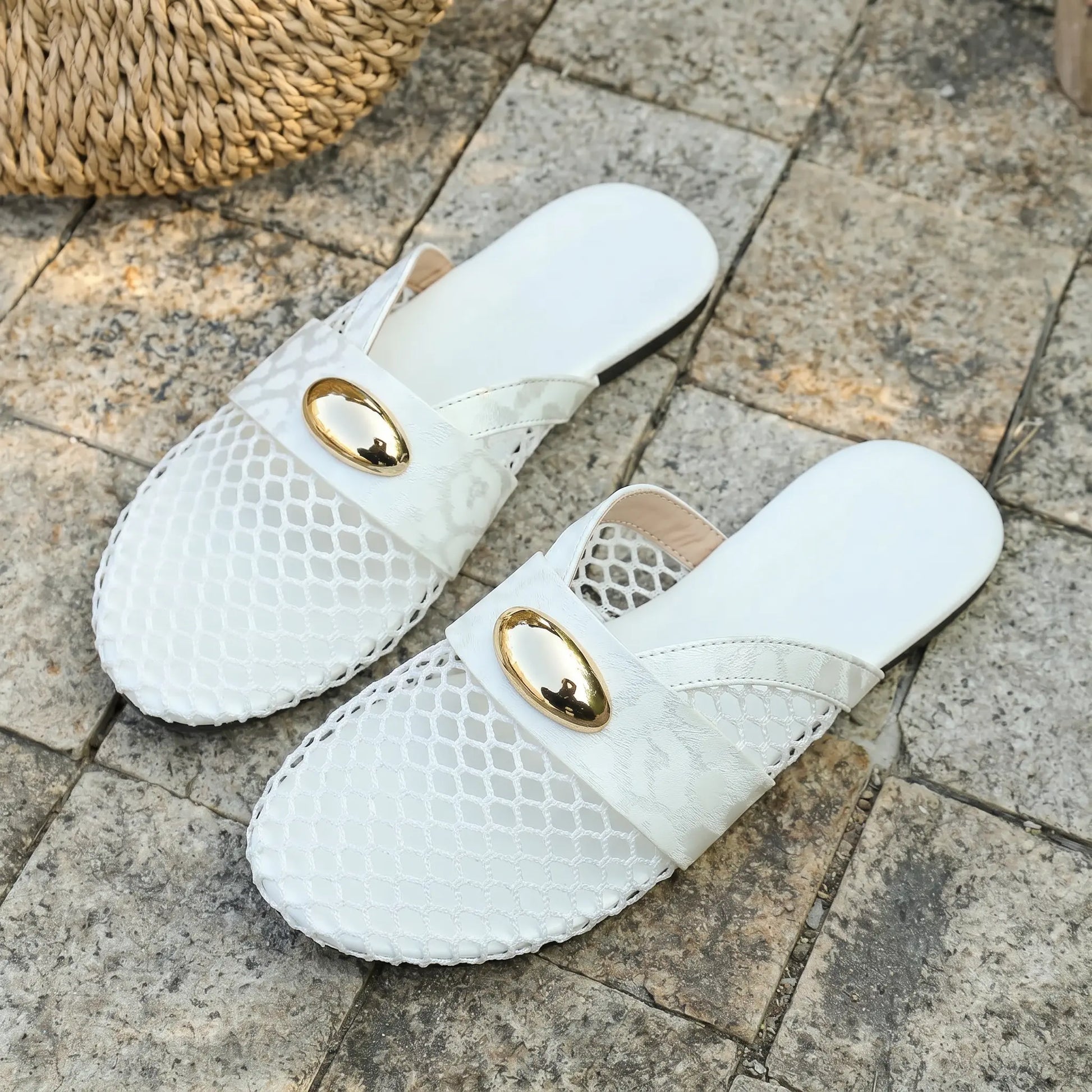 Pair of white Kryxos mesh pointed-toe flat mules with gold oval hardware and faux leather strap, presented on a textured stone surface with a woven basket accent.