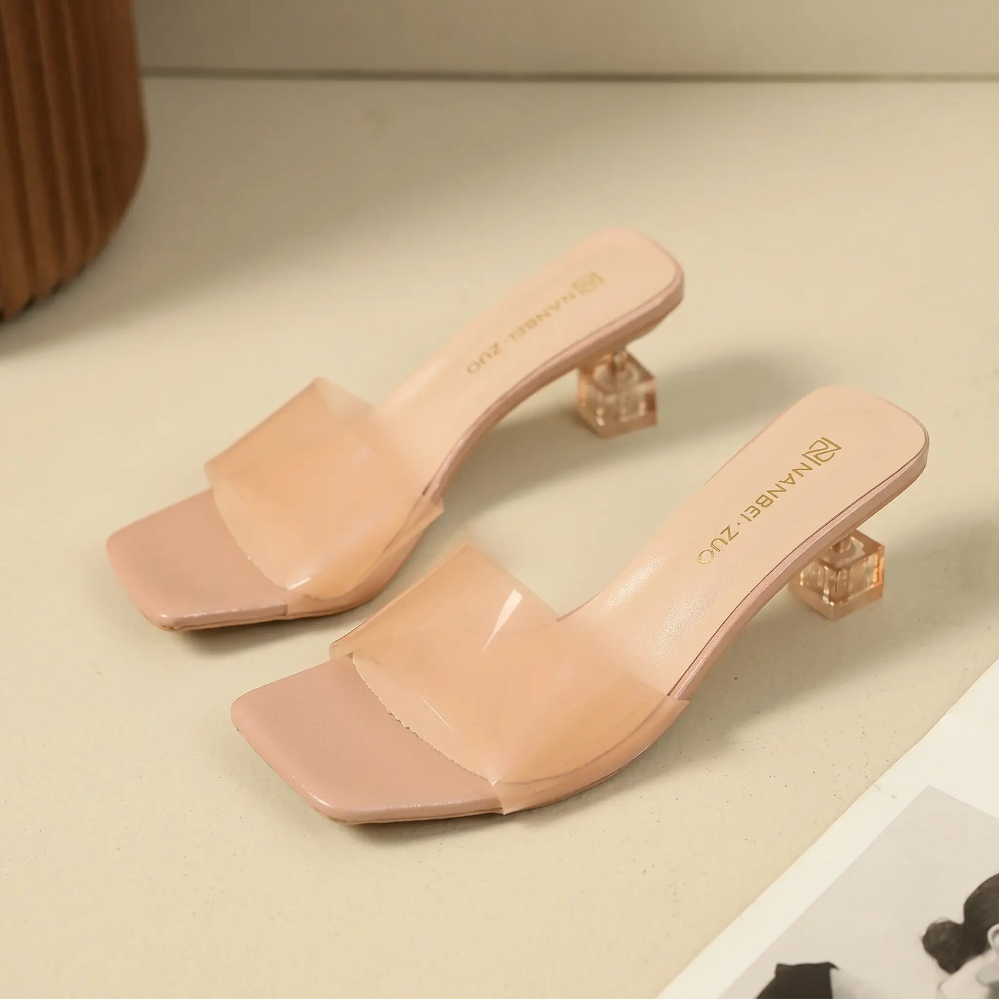 Nude pink slip-on mule sandals featuring a square toe, a single semi-transparent frosted strap, and a clear geometric block heel.