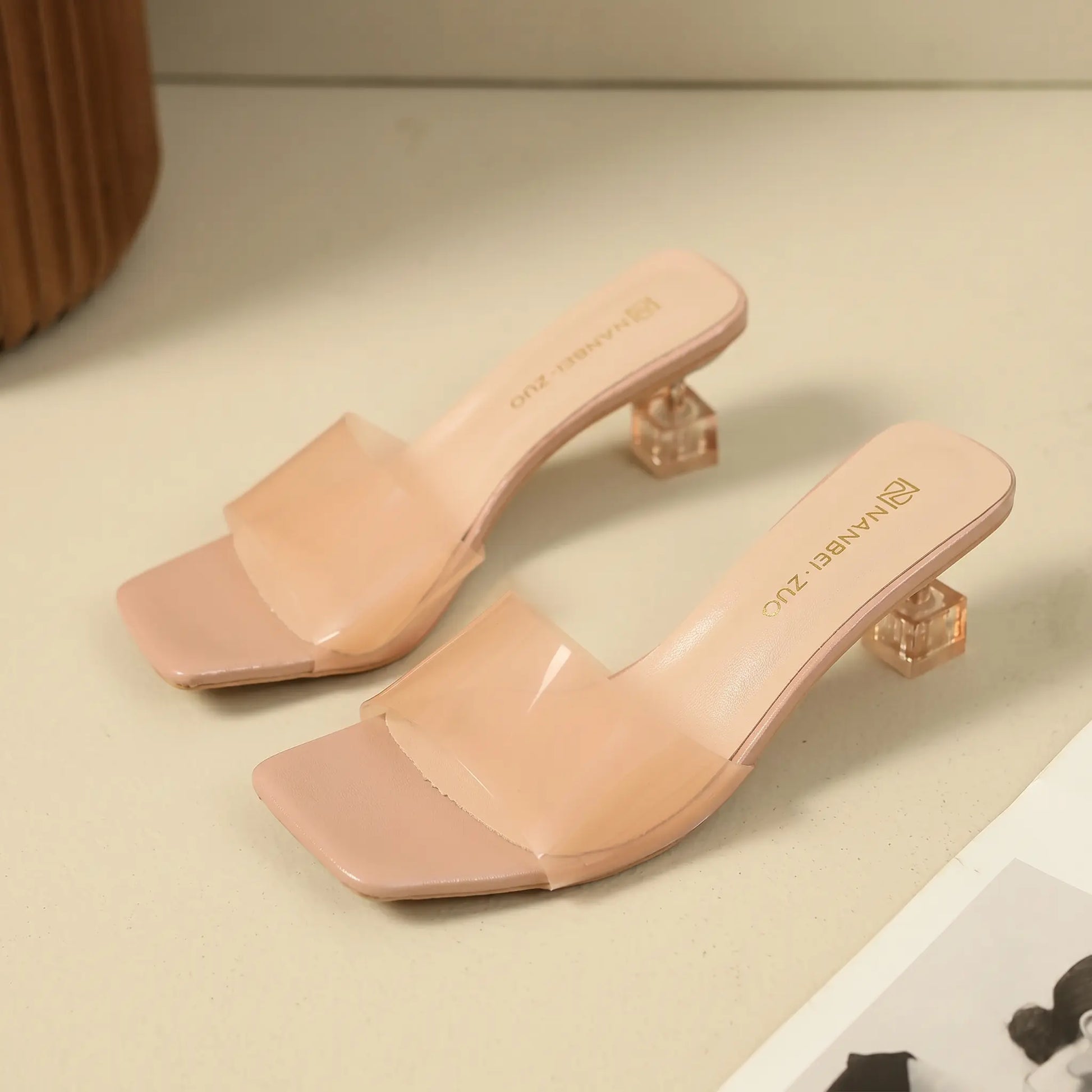 Nude pink slip-on mule sandals featuring a square toe, a single semi-transparent frosted strap, and a clear geometric block heel.