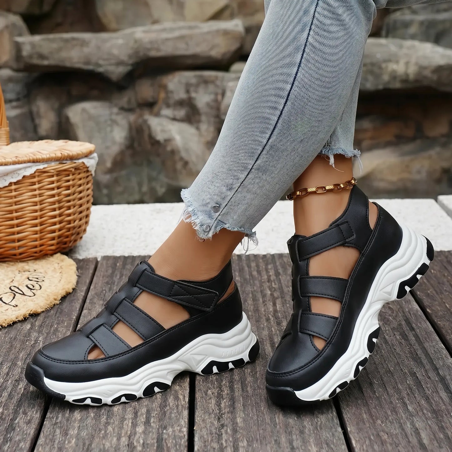 Black Kryzen sporty chic chunky platform sandals shown on model's feet with distressed cuffed jeans and a gold anklet, styled against a rustic wooden plank and stone background.