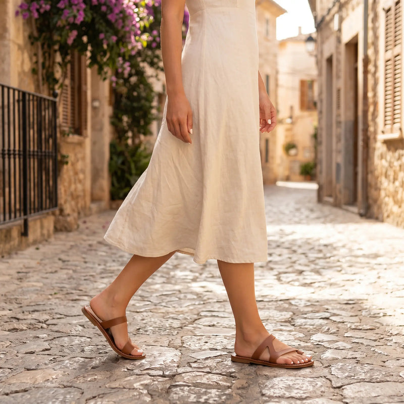 A woman wearing Kydra brown faux leather flat sandals with a toe-loop and asymmetrical strap walks on a charming cobblestone street, embodying effortless summer style.