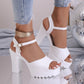 White Kylos platform high heel sandals worn by a model with distressed denim jeans, showcasing the fish-mouth peep-toe, ankle strap with gold charm, and chunky platform sole on a reflective surface with disco ball elements.