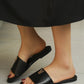 Close-up view of a woman's feet wearing black Kyluxa leather slide sandals with a metallic gold 'V' logo emblem on the strap, on a neutral tiled floor.