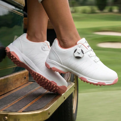 Women's Kymari white and blush pink golf shoes, featuring a rotary dial closure and aggressive spiked soles, worn by a person sitting on a golf cart with a green course visible in the background.