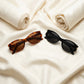 Two Kyndox premium acetate cat-eye sunglasses, one tortoiseshell and one piano black, with gold-tone accents, laid flat on elegant cream satin.