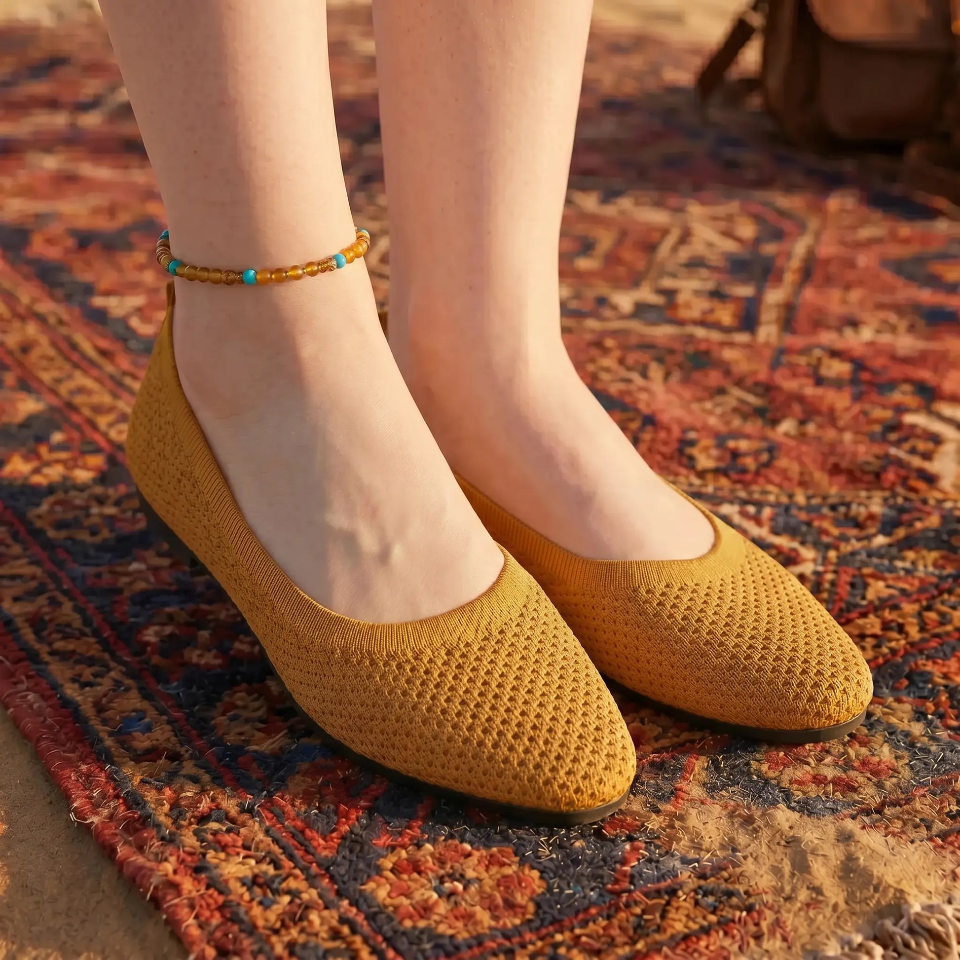Female model's feet wearing Kyneth mustard yellow breathable knit flats with a pointed toe and textured mesh upper, standing on a bohemian patterned rug. An ankle bracelet adorns one ankle.