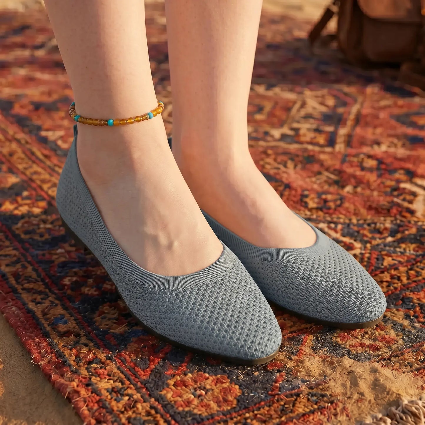 A pair of Kyneth slate grey knitted mesh pointed flats worn on feet with an anklet, standing on an ornate bohemian rug outdoors.