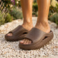 Person wearing Kynso Deep Coffee Brown platform lounge slides on a gravel path. The thick molded EVA sandals feature a wide strap, open toe, and architectural side cutout.