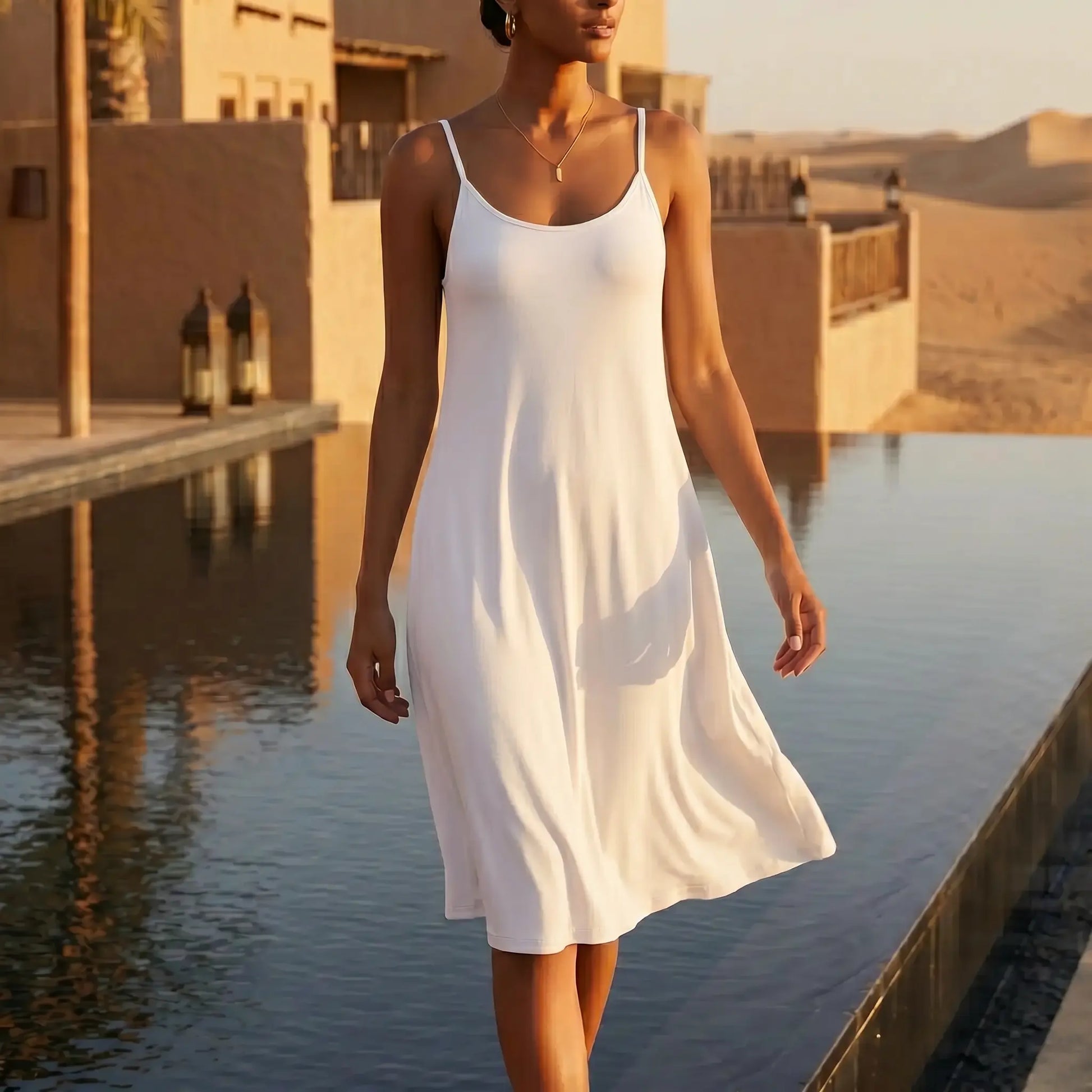 Woman wearing a Kynso premium white modal slip dress with thin spaghetti straps and a scoop neck, walking alongside a reflective pool with a desert architectural background at sunset.