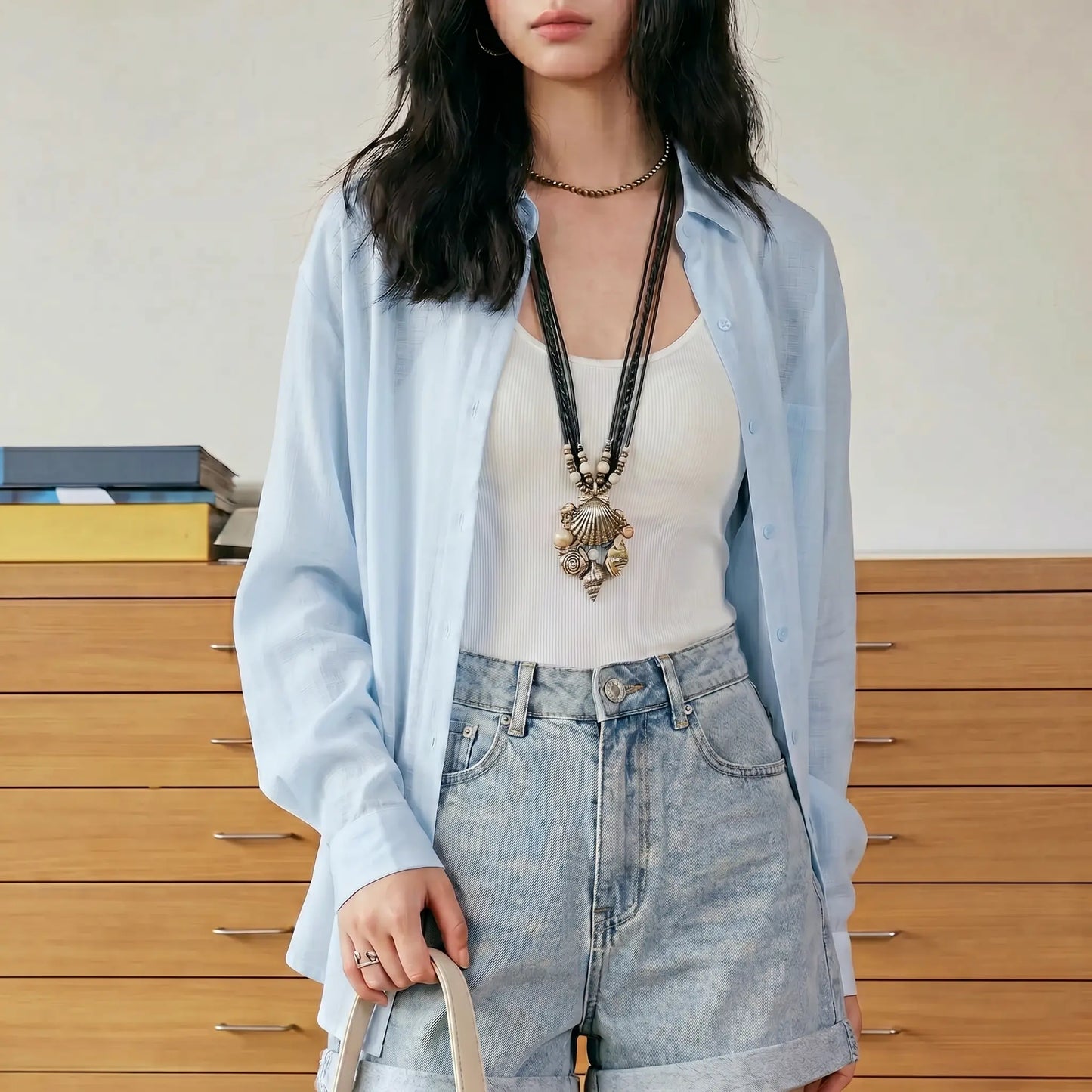 Female model wearing the Kynzor Breeze Light Blue Lightweight Tencel Button-Down Blouse, styled open over a white tank top with light wash denim shorts in a bright indoor setting.