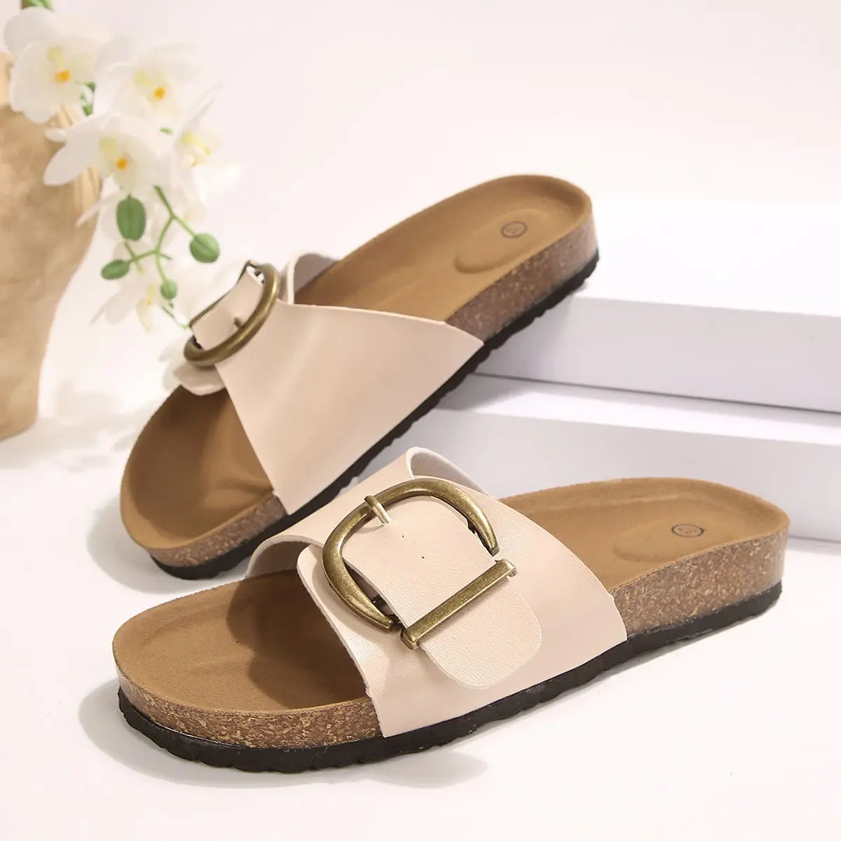 Pair of Kyraan women's beige comfort cork slides with antique brass buckle strap and open-toe design, displayed on a clean studio surface.