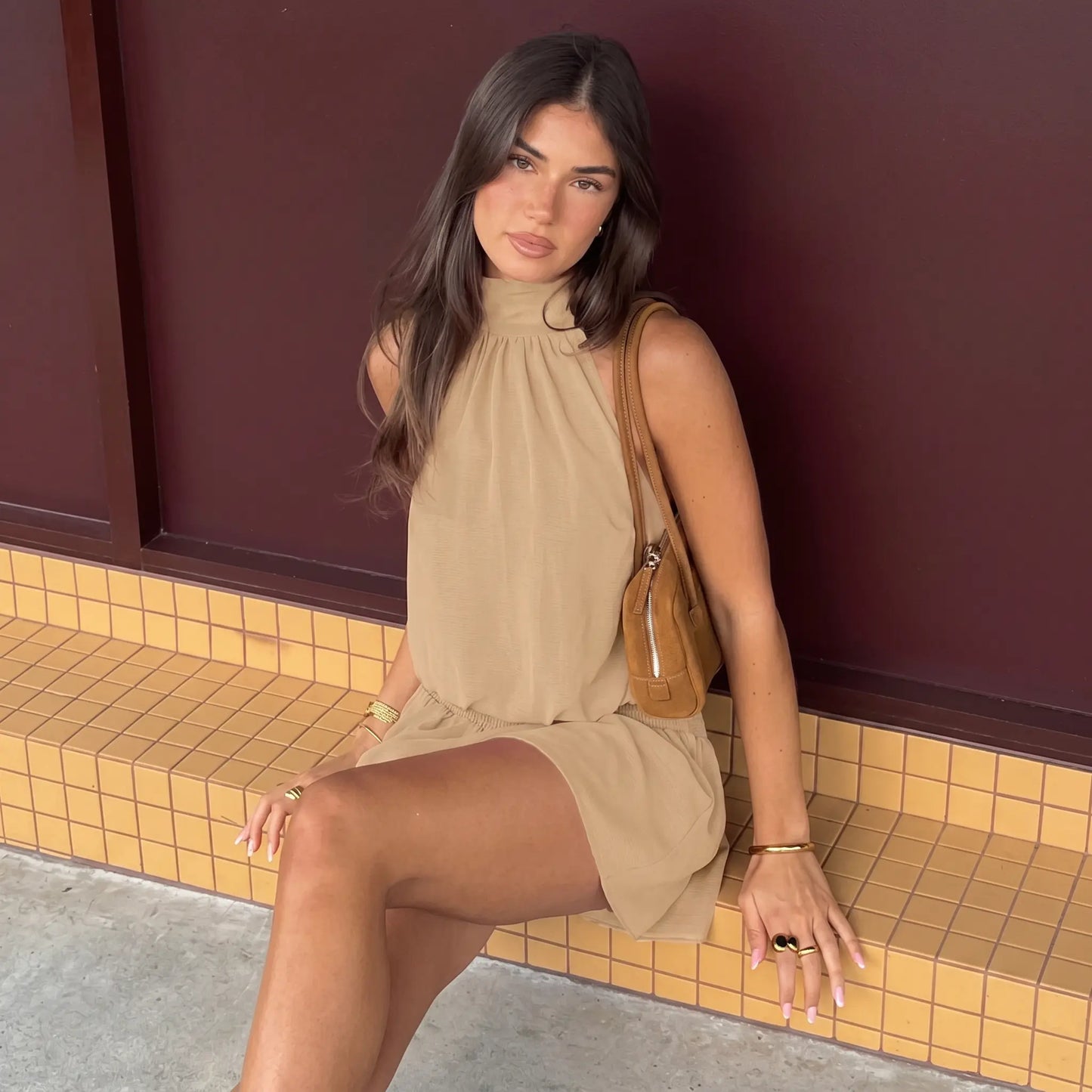 A woman wears a taupe halter neck mini dress with a smocked waist and flowy skirt, accessorized with a brown shoulder bag and jewelry, seated outdoors.
