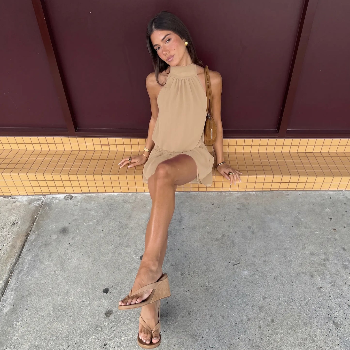 A woman sits outdoors wearing a taupe halter neck mini dress with a smocked waist and flowy skirt, styled with a small brown bag and wedge sandals.