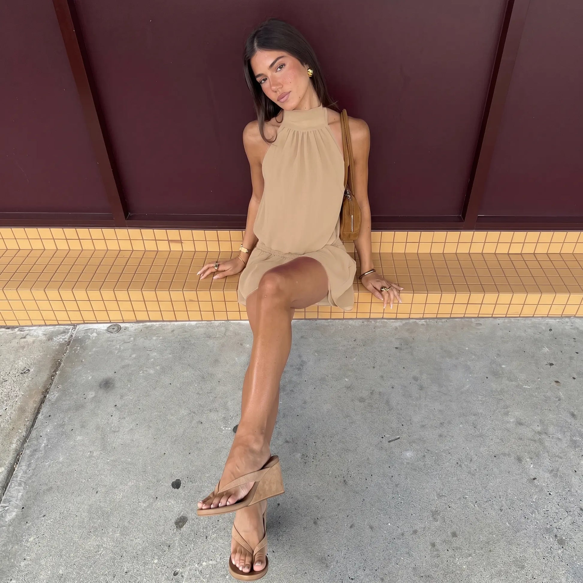 A woman sits outdoors wearing a taupe halter neck mini dress with a smocked waist and flowy skirt, styled with a small brown bag and wedge sandals.
