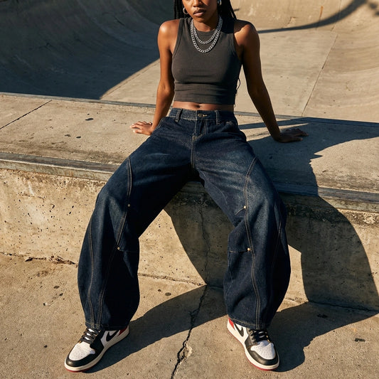 Kyro architectural curved leg jeans in midnight indigo, worn by a model sitting on concrete to showcase the voluminous balloon fit and front seam details.