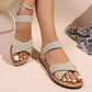 Woman's feet wearing elegant gold shimmering embellished flat sandals with crossover elasticated straps and a delicate pearl anklet against a soft neutral background.