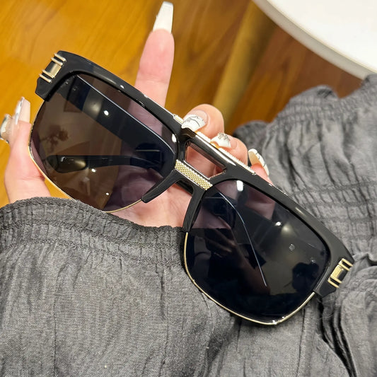 Oversized black square sunglasses with gold double bridge and gradient grey lenses, held at a three-quarter angle. Detailed gold hardware on temples.