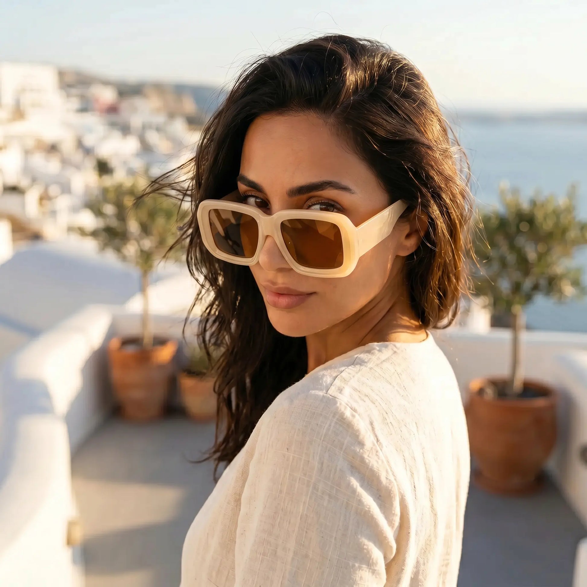 A woman wears Kyzeth oversized square sunglasses with thick beige frames and amber lenses, looking back over her shoulder in a sunny Mediterranean landscape.