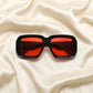 Kyzeth oversized square statement sunglasses featuring high-gloss black frames and vibrant red lenses, elegantly displayed on a draped cream satin fabric.