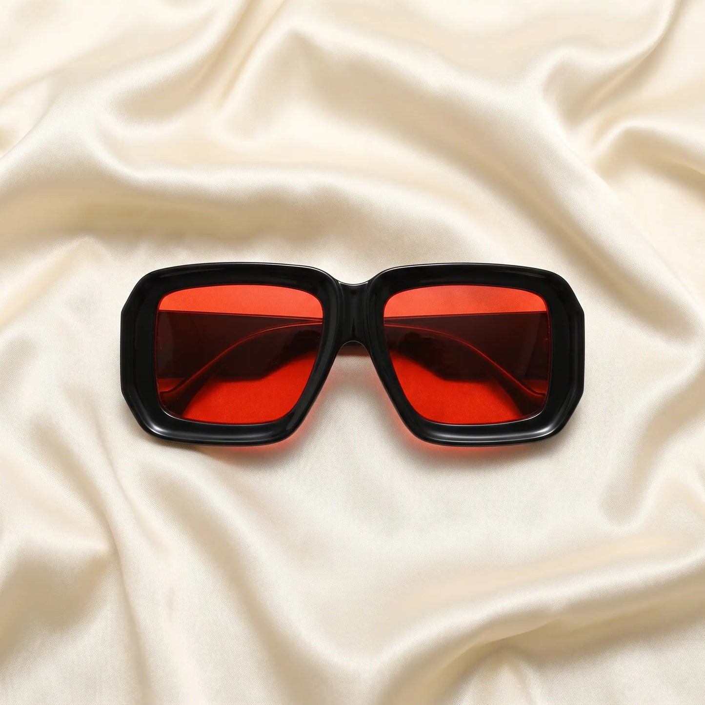 Kyzeth oversized square statement sunglasses featuring high-gloss black frames and vibrant red lenses, elegantly displayed on a draped cream satin fabric.