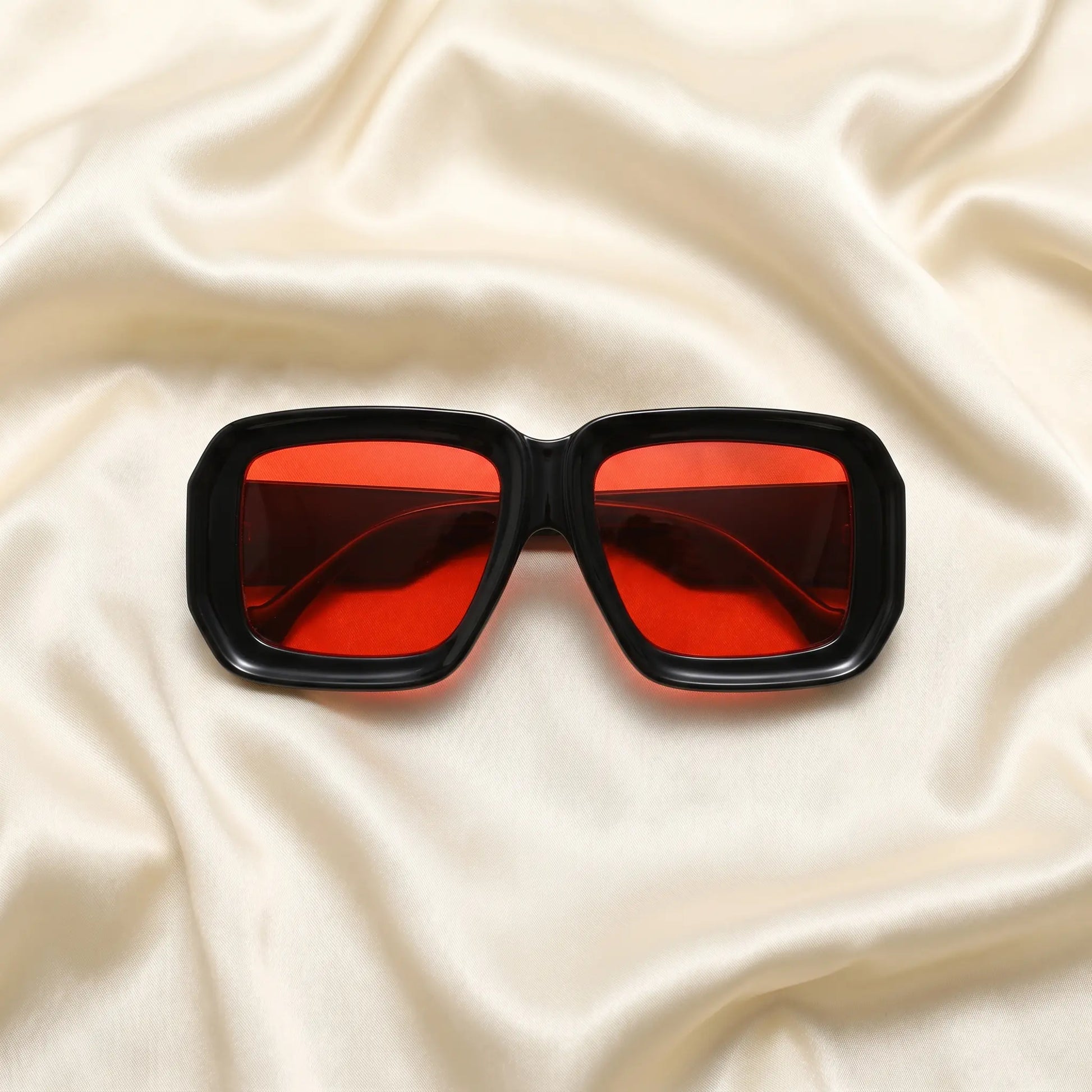 Kyzeth oversized square statement sunglasses featuring high-gloss black frames and vibrant red lenses, elegantly displayed on a draped cream satin fabric.