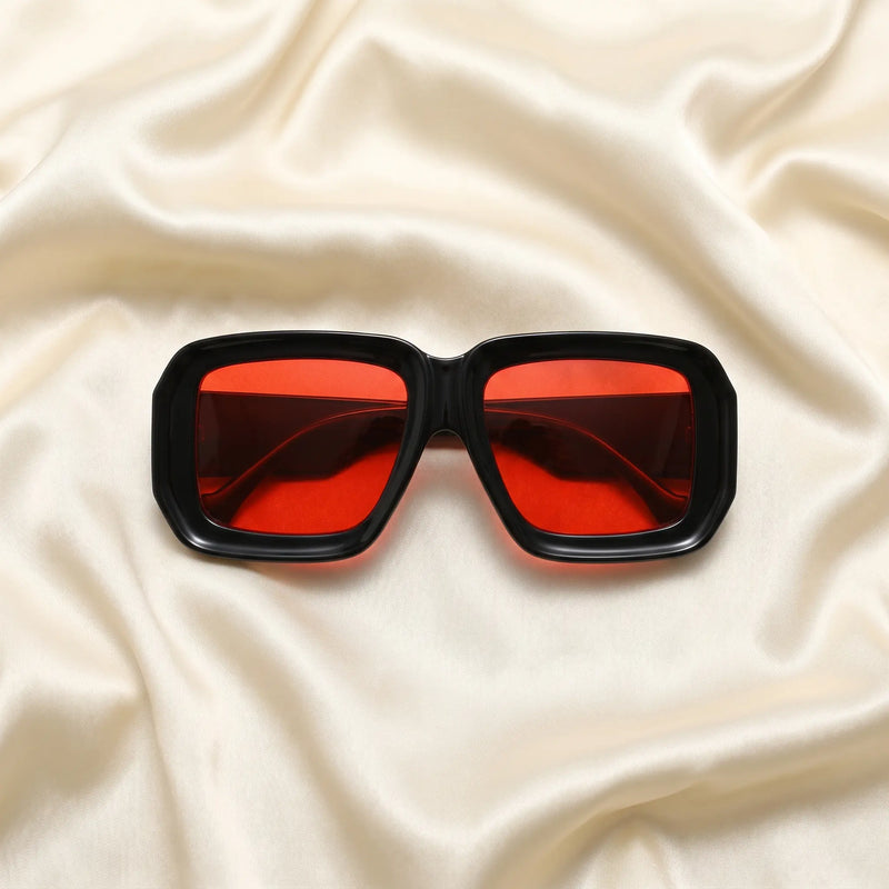 Kyzeth oversized square statement sunglasses featuring high-gloss black frames and vibrant red lenses, elegantly displayed on a draped cream satin fabric.