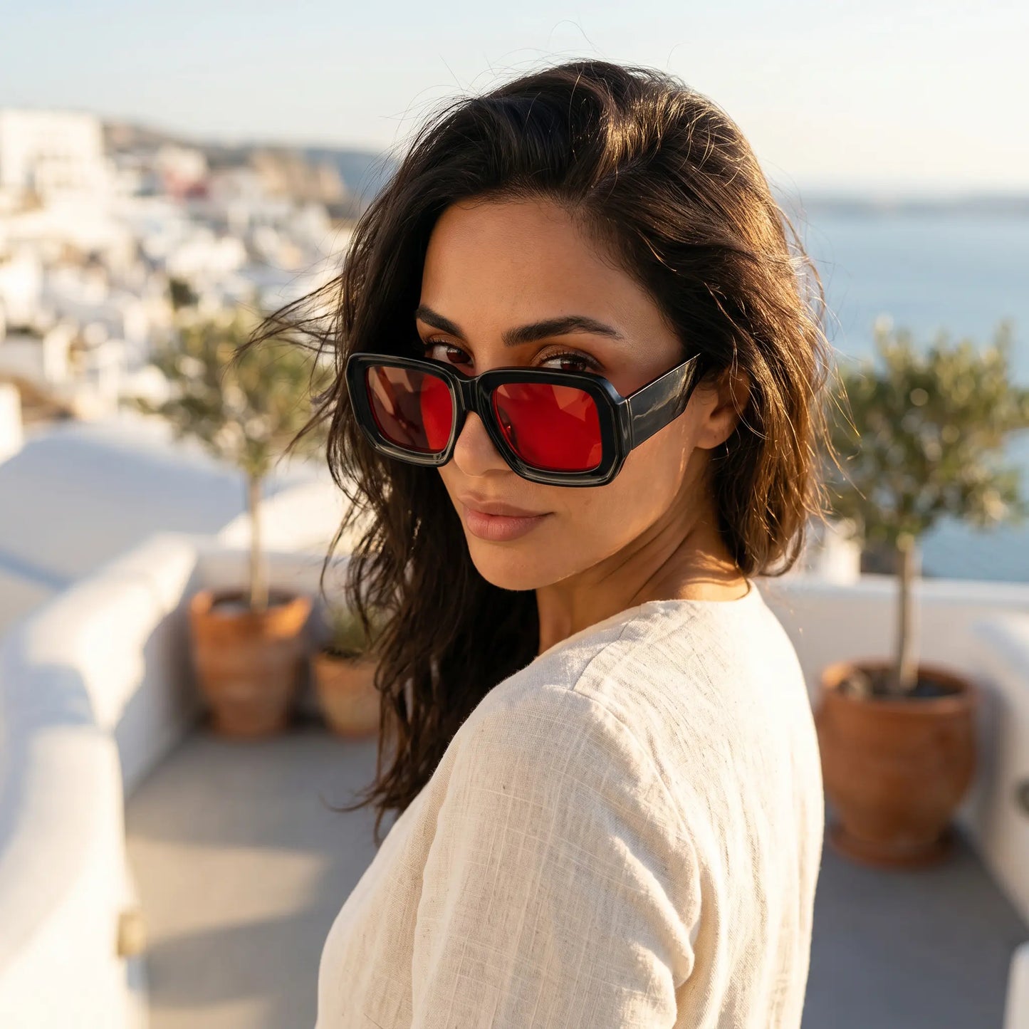 A woman wears Kyzeth oversized black square statement sunglasses with vibrant red lenses, looking over her shoulder on a sunny balcony overlooking a Mediterranean town.