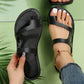 Black asymmetrical open-toe faux leather slide sandals worn on a foot, featuring white contrast stitching on the flat sole and a delicate butterfly anklet.