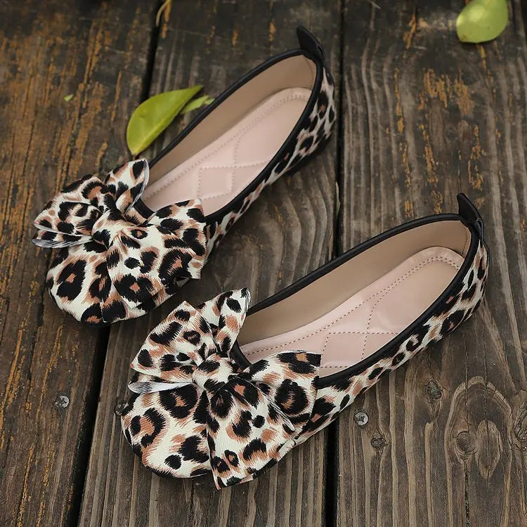 Kaeluxe leopard print ballet flats with a prominent layered bow detail, viewed from a flat lay angle on a rustic wooden surface.