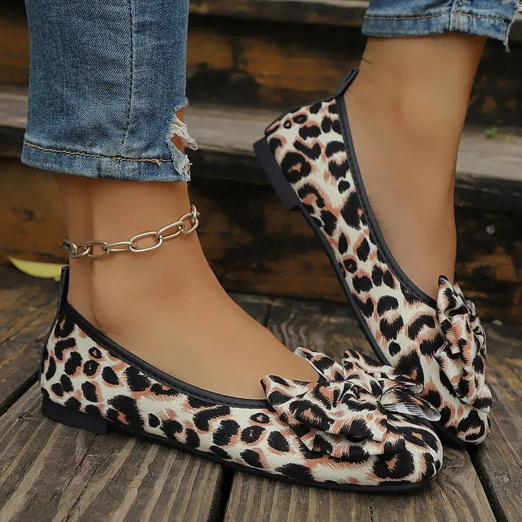 Women's leopard print ballet flats featuring a large layered bow detail, worn with ripped jeans and a silver anklet on a wooden surface.