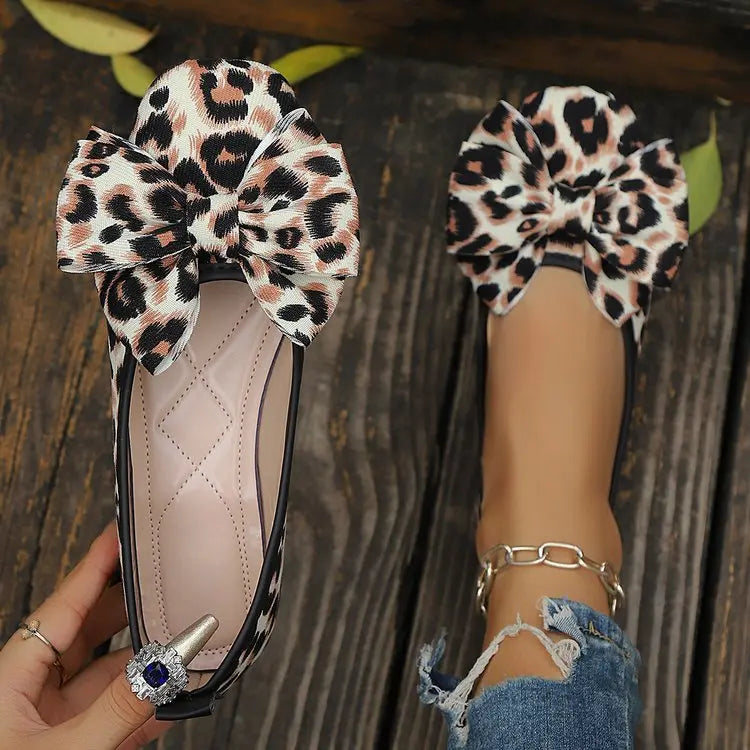 Women's leopard print ballet flats with large layered bows on a wooden surface, shown on a model wearing ripped jeans and a silver anklet.