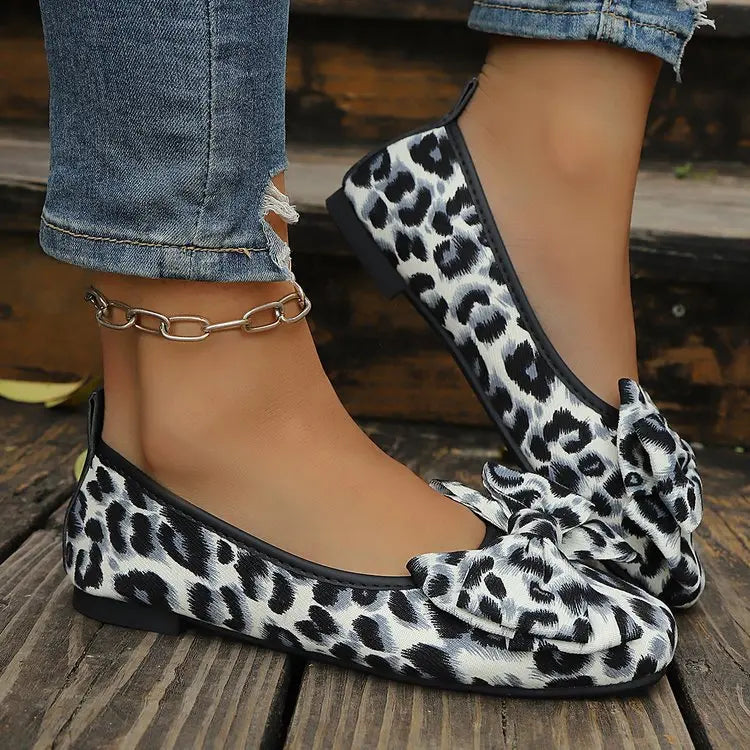 Women's leopard print ballet flats with a prominent layered bow, styled with ripped denim jeans and a chain anklet, shown on wooden steps.