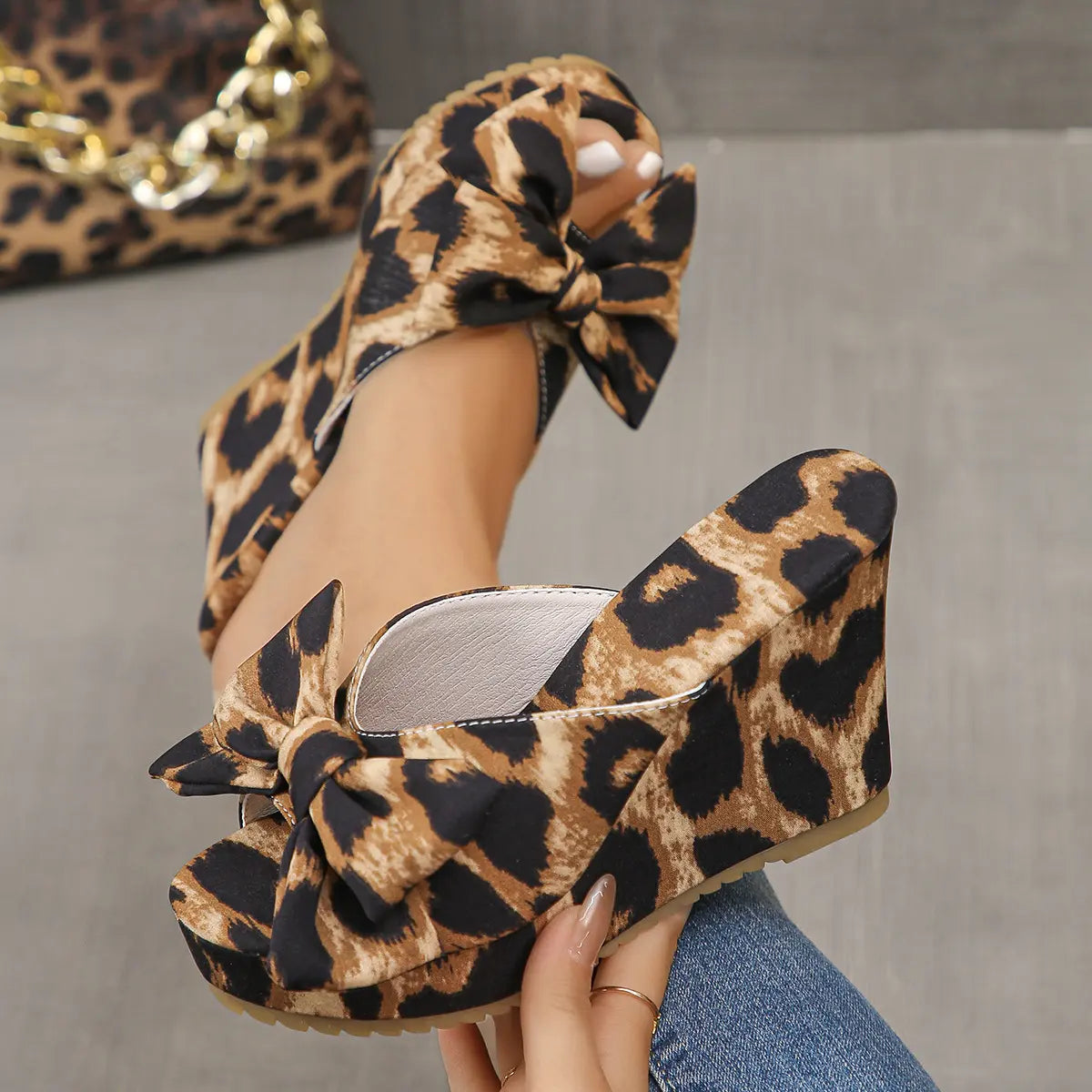 Close-up of leopard print wedge sandals with oversized bows, worn on a woman's feet, showcasing the peep-toe and platform heel on a grey studio background.