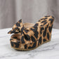 Leopard print wedge platform sandals featuring an oversized bow embellishment and peep-toe design, shown from a three-quarter angle on a marble surface.
