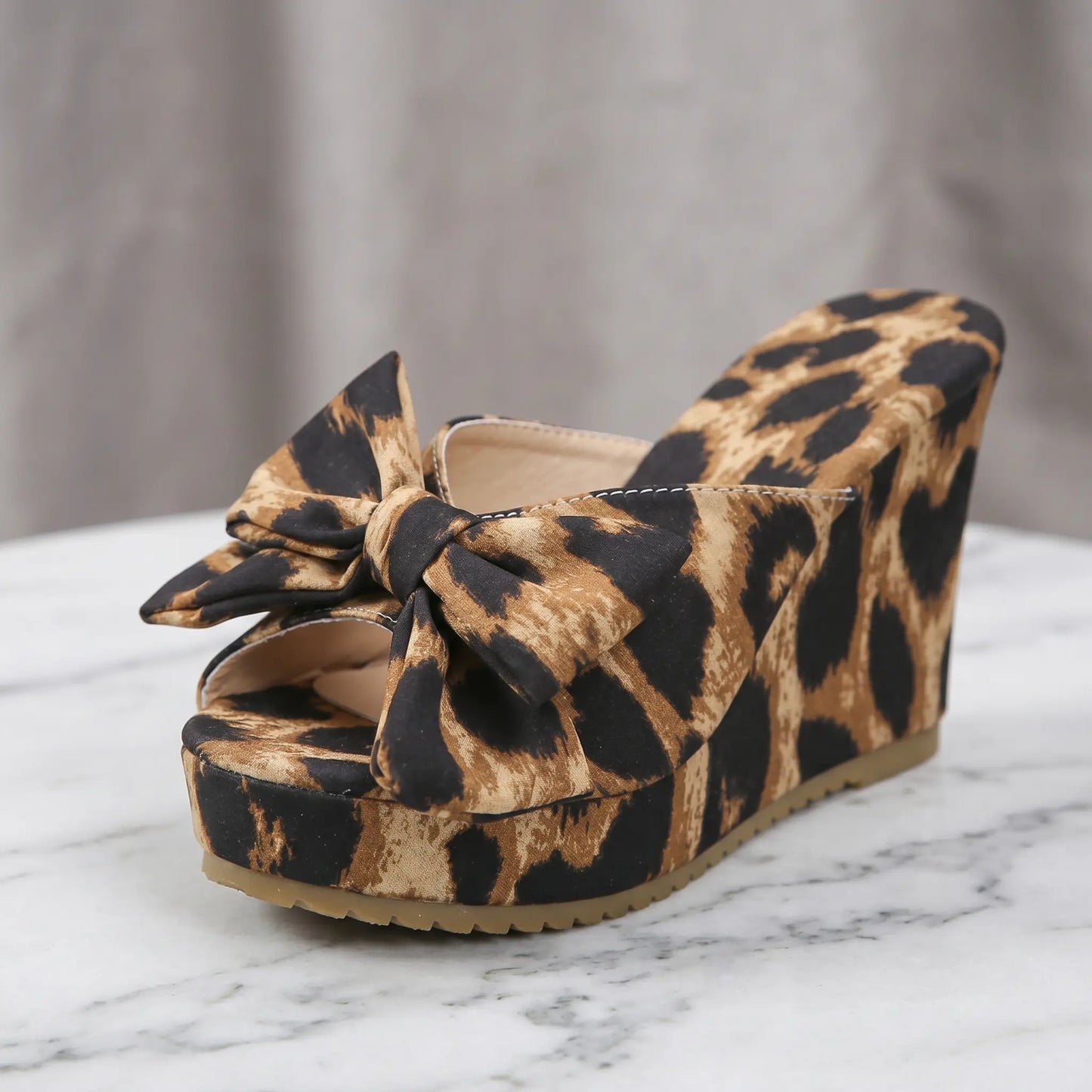 Leopard print wedge platform sandals featuring an oversized bow embellishment and peep-toe design, shown from a three-quarter angle on a marble surface.