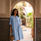 Woman wearing a light blue crinkled cotton linen V-neck midi dress with half sleeves and front patch pockets, styled in a Moroccan courtyard.
