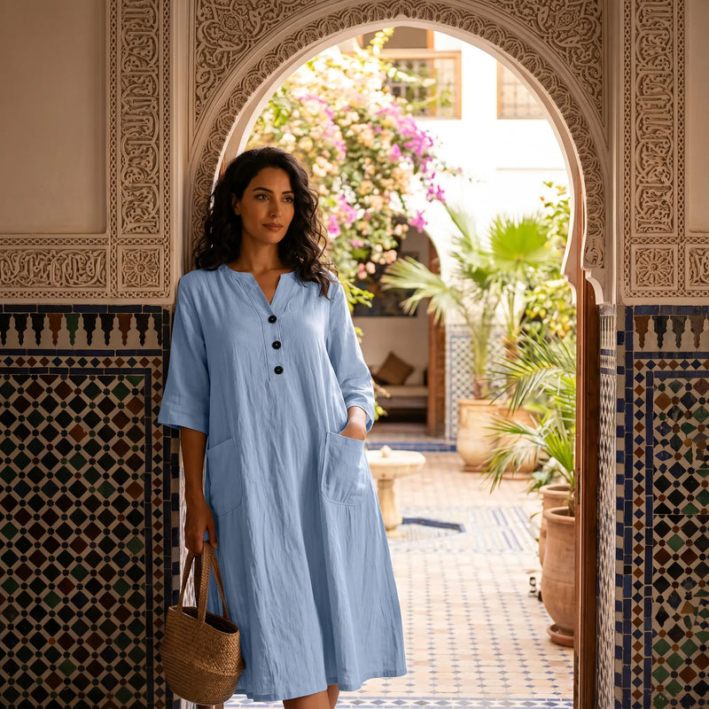 Woman wearing a light blue crinkled cotton linen V-neck midi dress with half sleeves and front patch pockets, styled in a Moroccan courtyard.