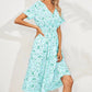 A woman models a light blue V-neck midi dress with a delicate ditsy floral print, short flutter sleeves, and a cinched smocked waist, shown in a three-quarter view against a textured wall.
